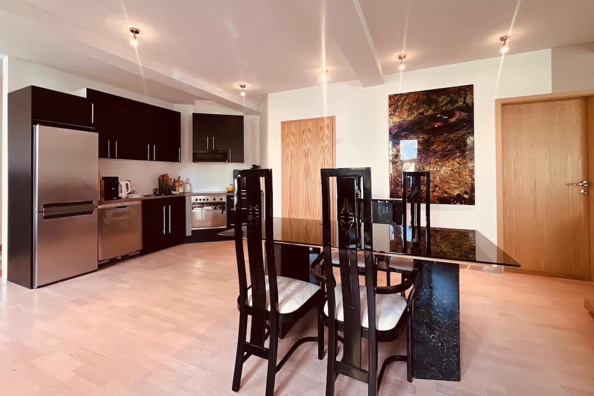 A modern kitchen with dark cabinets, a stainless steel refrigerator, a stove, and a dining area with a black table and chairs.
