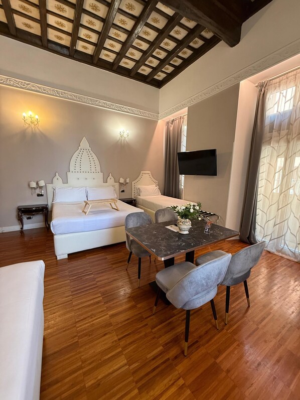 A bedroom with a large bed, a dining table with chairs, a TV mounted on the wall, and a chandelier.