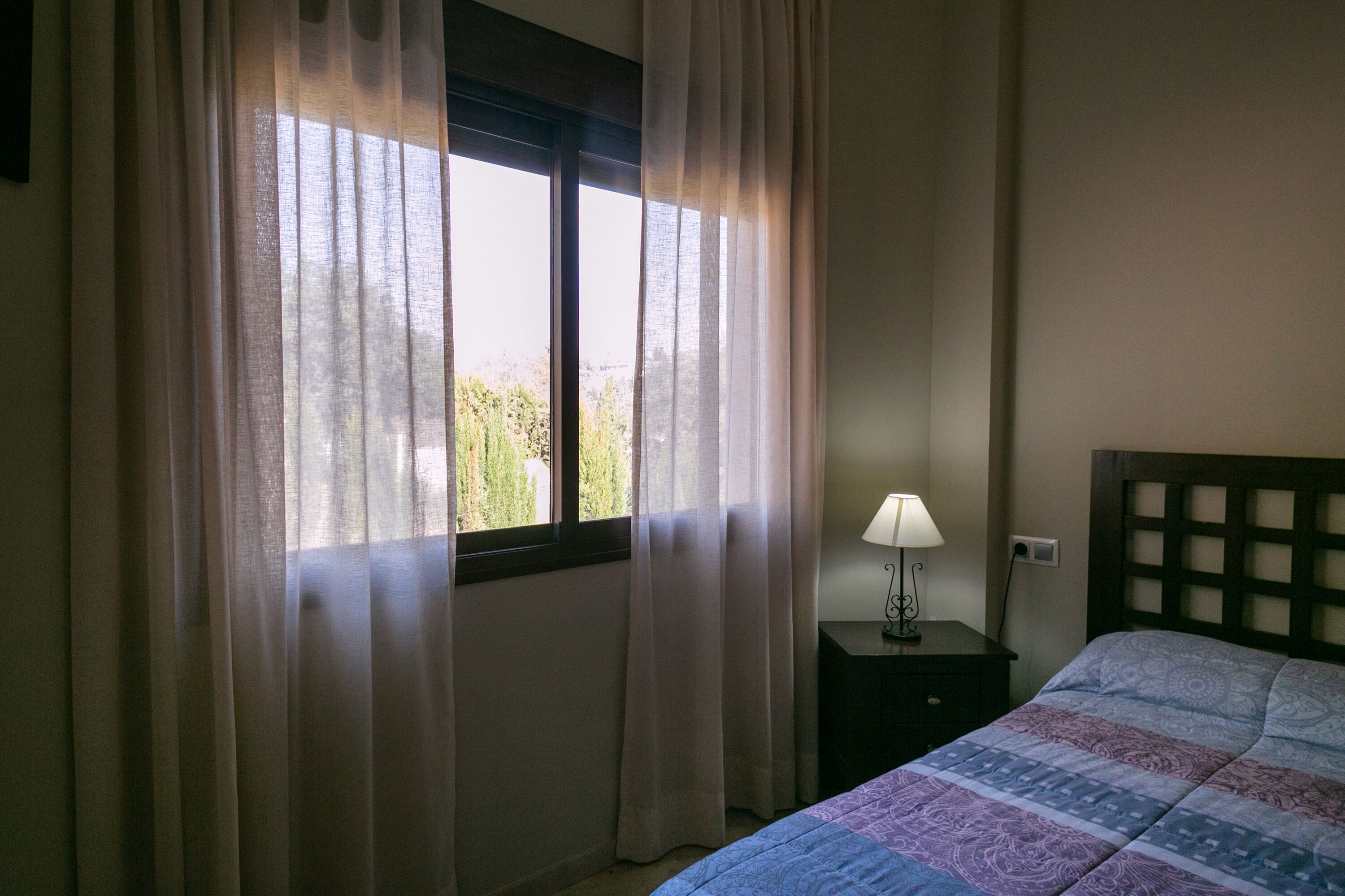 A bedroom with a bed, a nightstand with a lamp, and a window with sheer curtains.