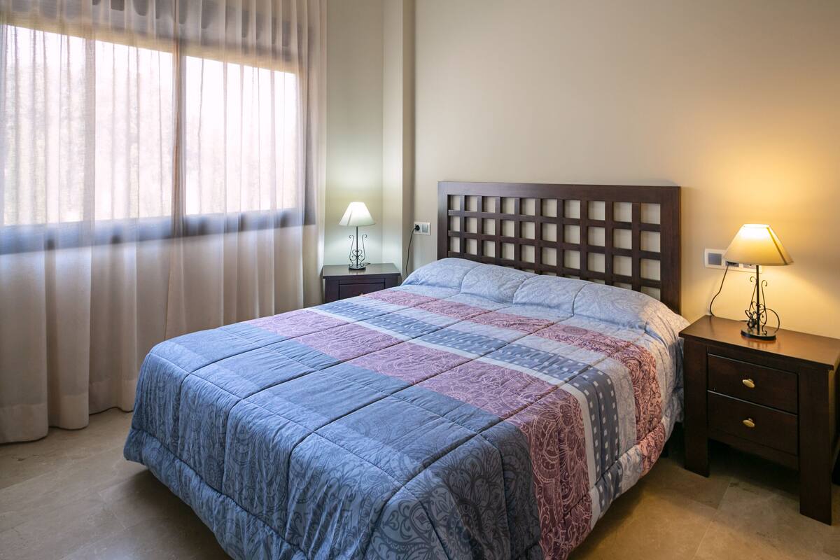 Un lit bien fait, recouvert d’une couvre-lit à motifs, avec une tête de lit en bois et deux tables de chevet dotées de lampes.