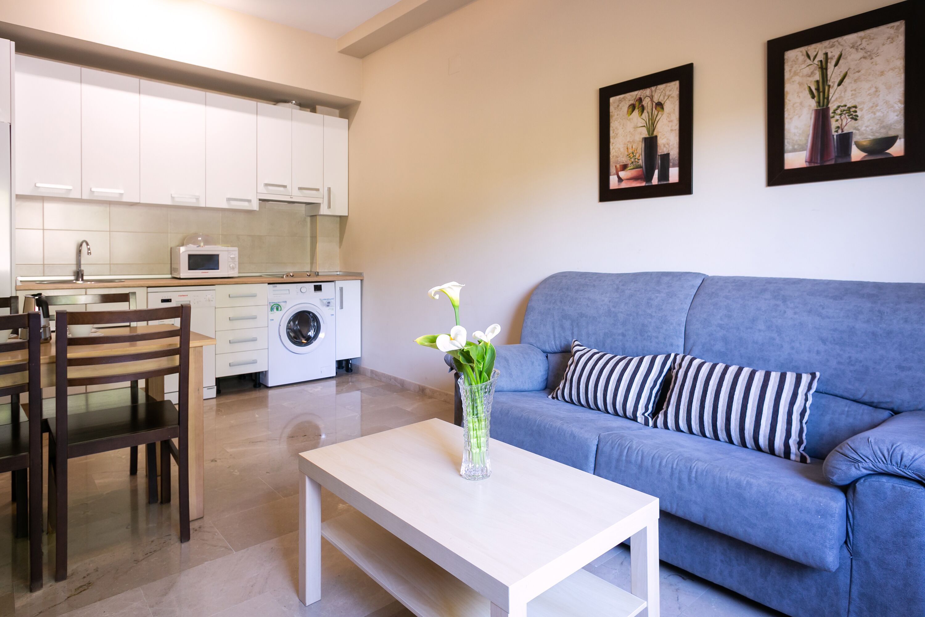 Una sala de estar moderna con un sofá azul, una mesa de comedor de madera y una mesa de centro blanca. Se observa una lavadora al fondo, y dos cuadros en la pared.