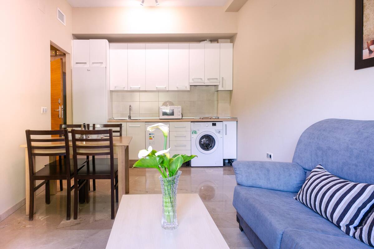 Un espace de vie compact comprenant un coin repas, un canapé, une table basse avec un vase, un lave-linge et un four à micro-ondes.