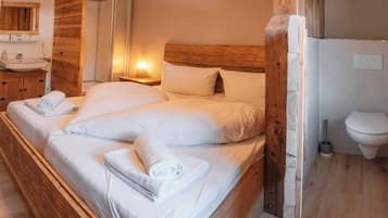 Een slaapkamer met een houten hoofdbord, een houten kledingkast, een nachtkastje met een lamp, en een badkamer met toilet en wastafel.