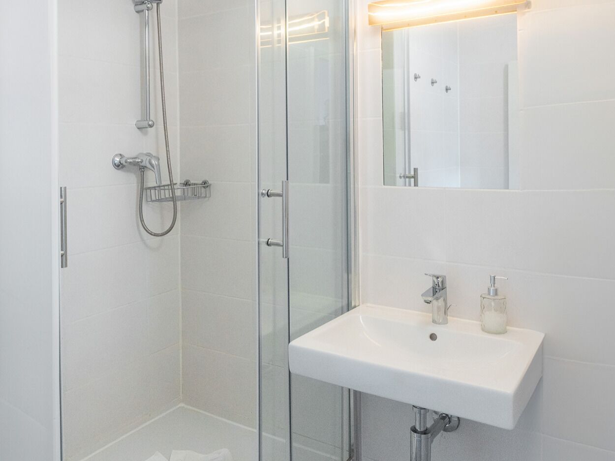 A modern bathroom with a glass shower enclosure, a white sink with a soap dispenser, and a mirror above the sink.