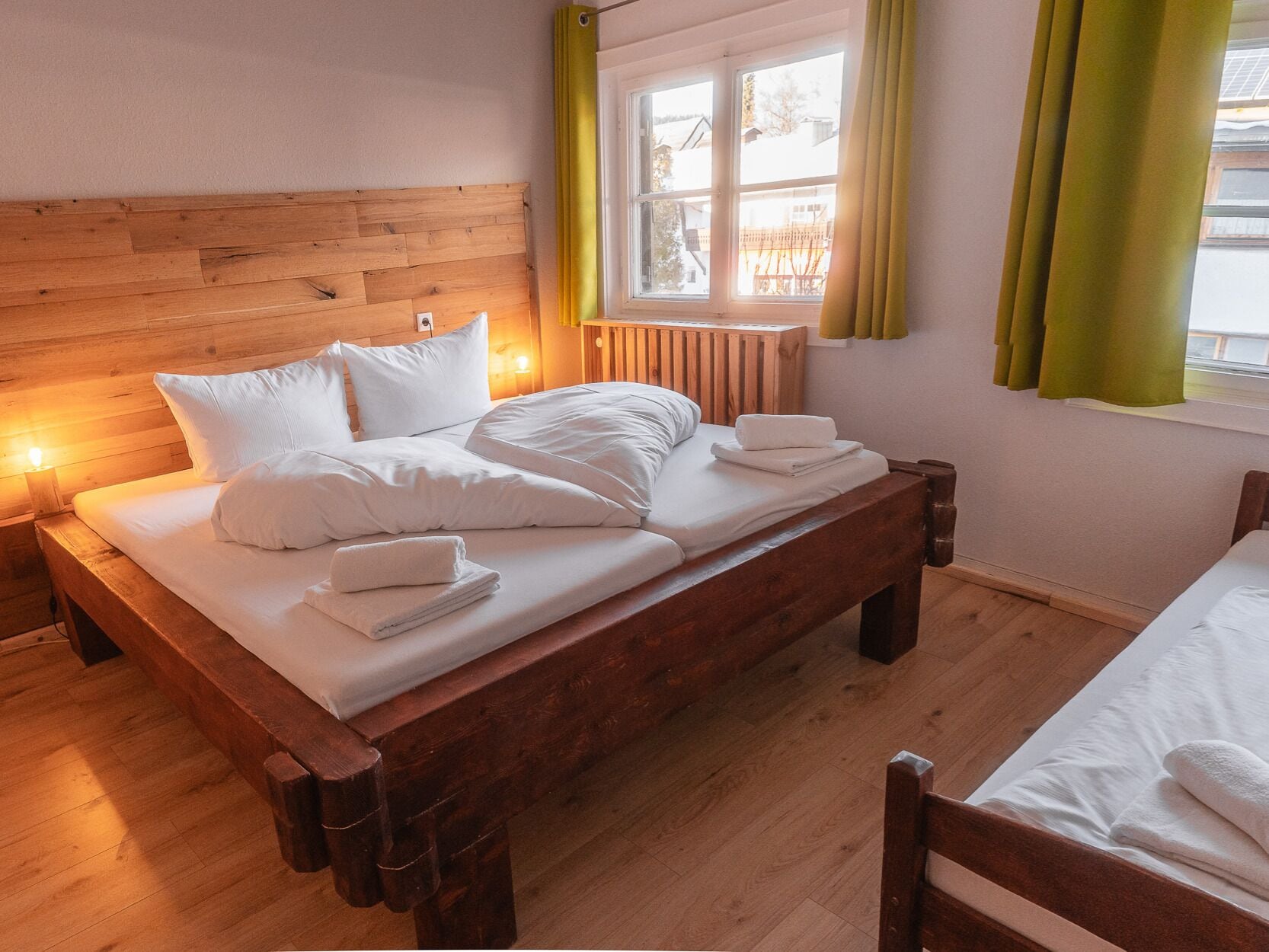 Una cama de madera con sábanas y almohadas blancas, una mesita de noche con lámpara, y una ventana con cortinas.