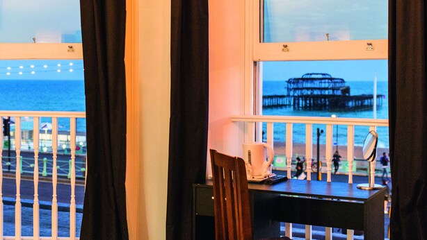 A hotel room with a bed, a desk, and a chair. There is a view of the beach and pier through the window. - West Beach Hotel (Brighton)