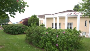 A building with a beige facade and white columns, surrounded by well-maintained hedges and a grassy lawn with scattered flowers. - Hôtel Relais d'Etretat (Bolbec)