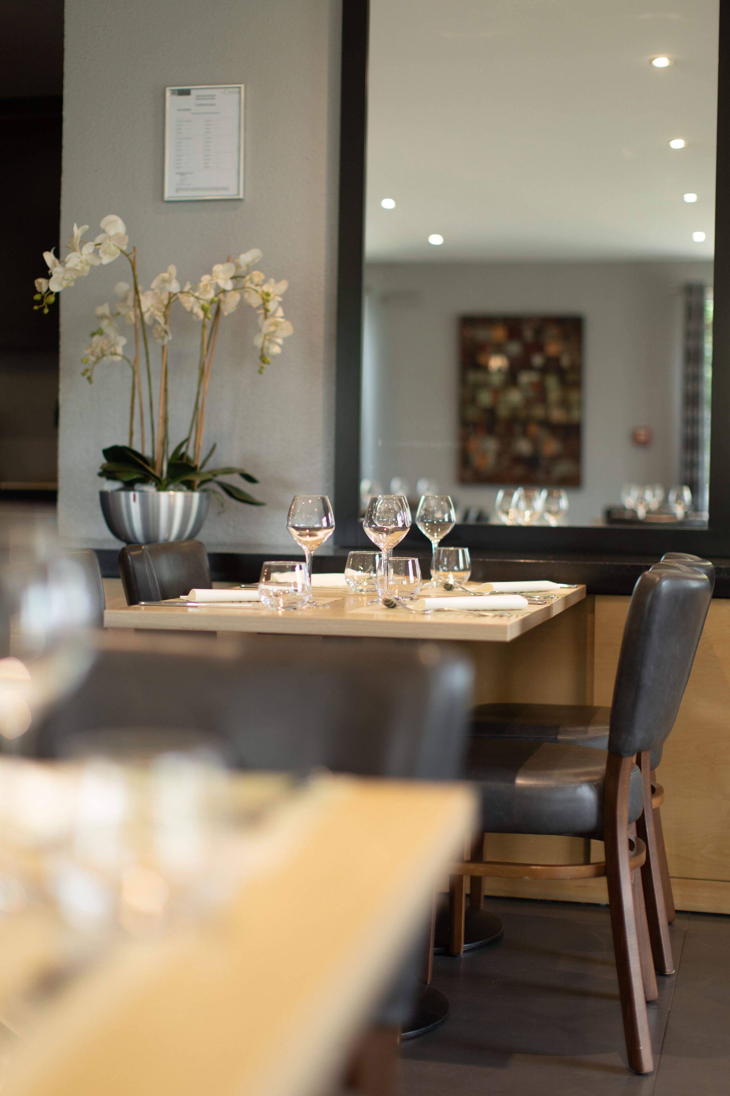 Un coin repas avec une table dressée pour deux personnes, un pot d’orchidée et un menu encadré accroché au mur.