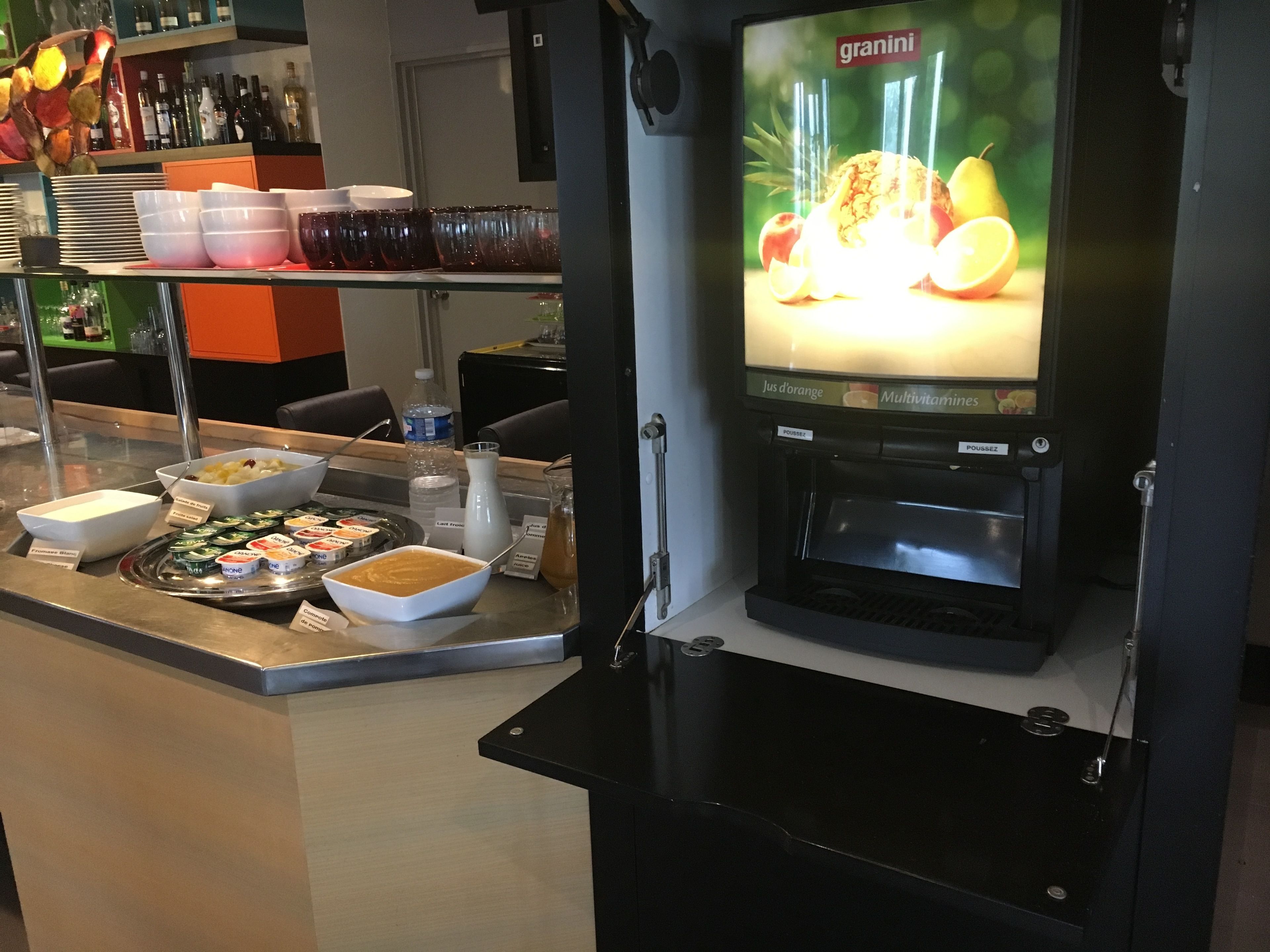 Una barra de desayuno con diversos alimentos y una máquina Granini que muestra imágenes de frutas.