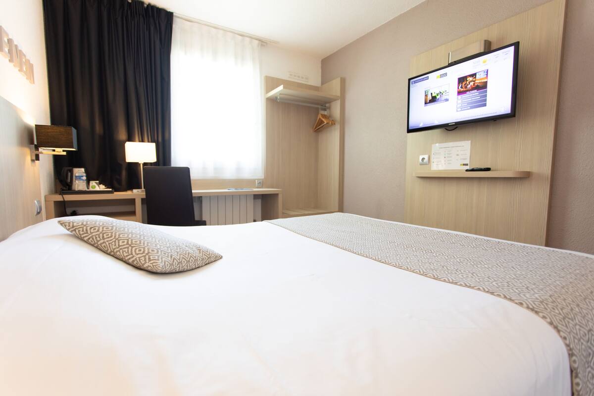 A hotel room with a large bed, a desk with a computer, a TV mounted on the wall, and a window with curtains.