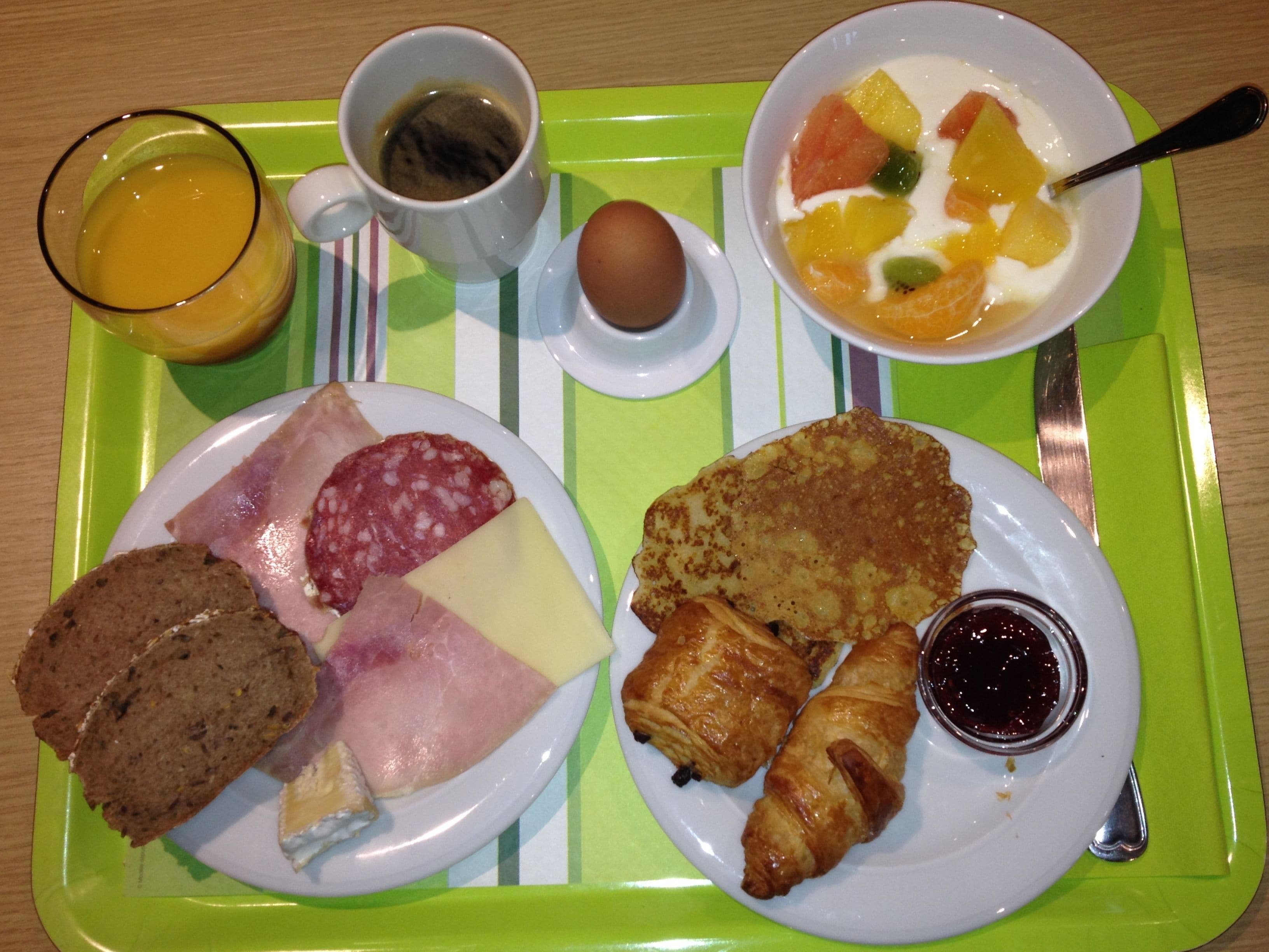 Bandeja de desayuno con jamón, queso, pan, croissants, un tazón de fruta, un vaso de jugo de naranja, una taza de café y un recipiente pequeño de mermelada.