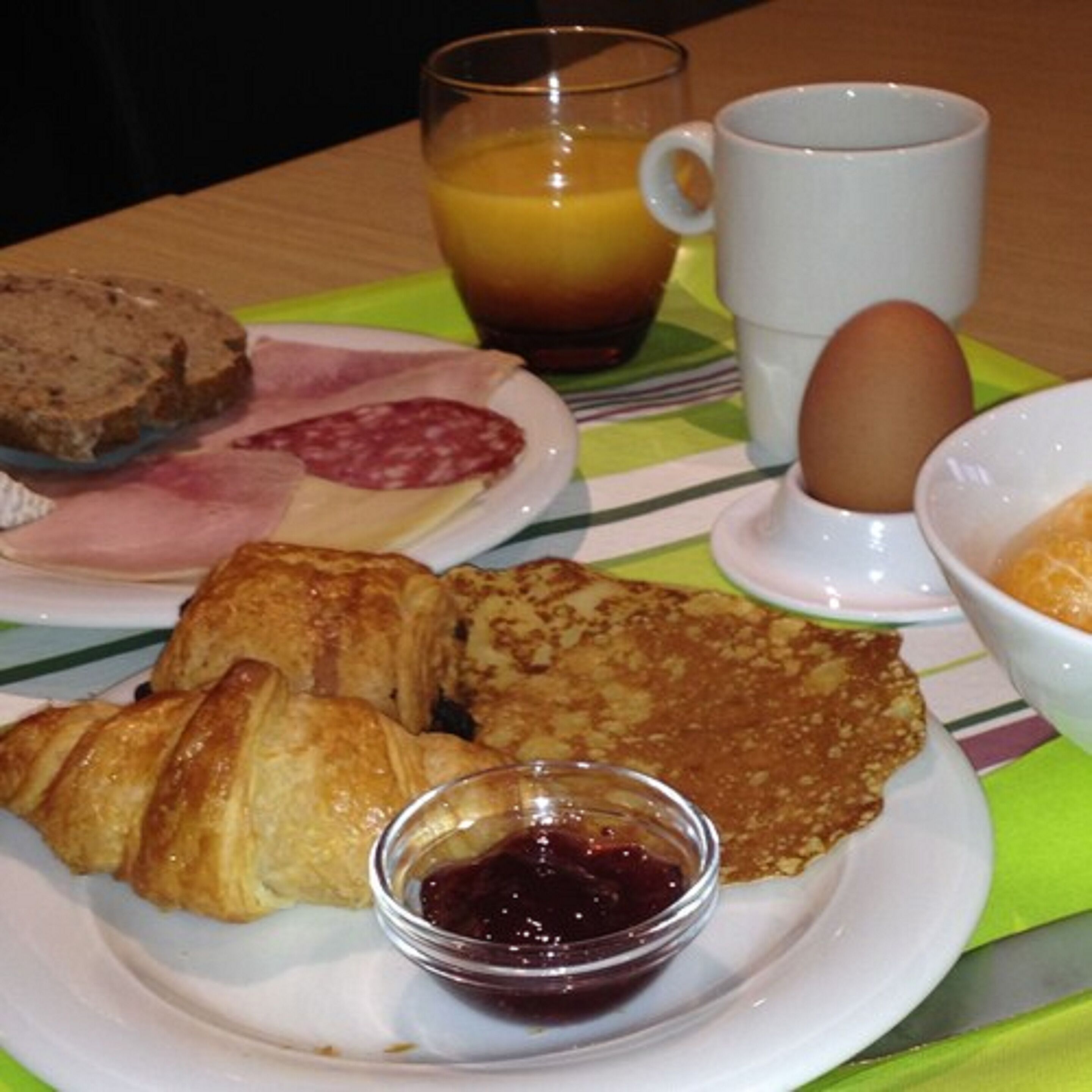 A breakfast plate with croissants, ham, a slice of bread, a glass of orange juice, a cup, an egg, and a small bowl of jam.
