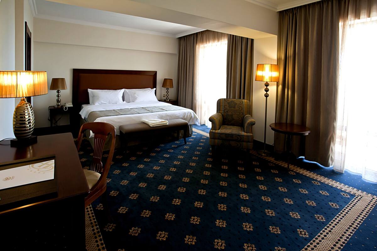 A hotel room with a large bed, a desk with a lamp, a chair, and a carpeted floor.