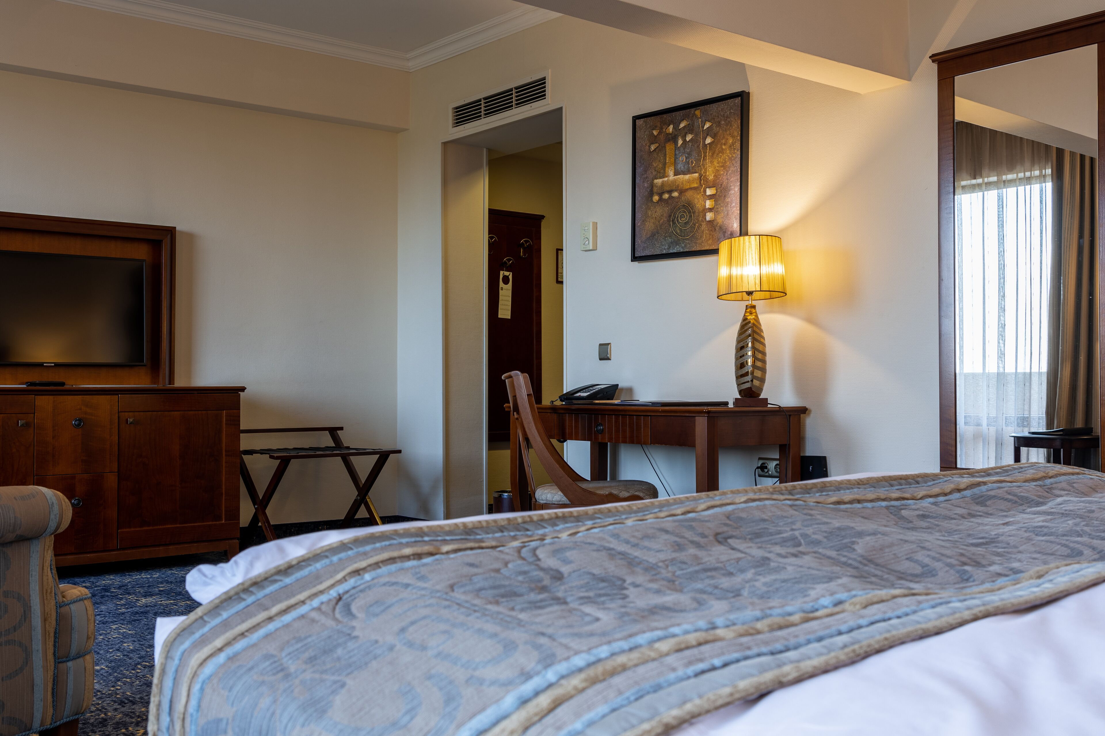 A hotel room with a large bed, a desk with a lamp, a chair, a television, and a door leading to another room.