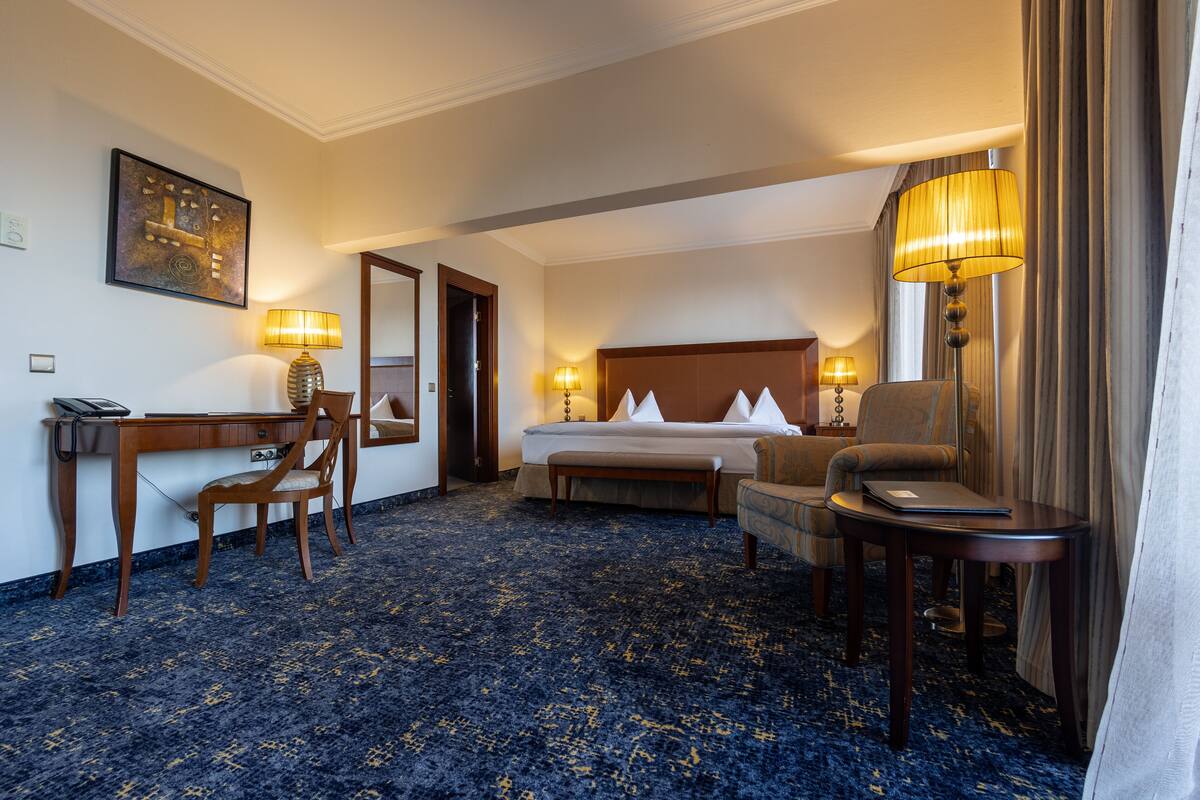 A hotel room with a bed, two bedside tables with lamps, a desk, a chair, and a carpeted floor.