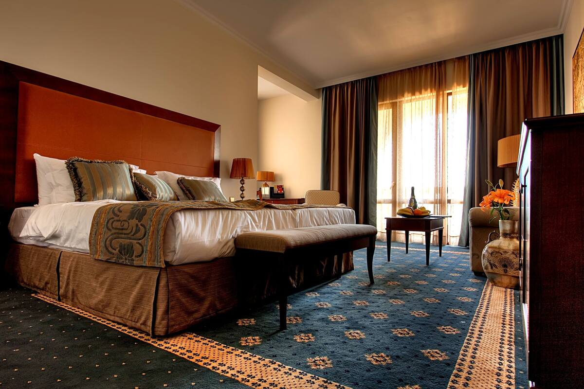 A hotel room with a large bed, a bench, a small table, a vase with flowers, and a lamp.