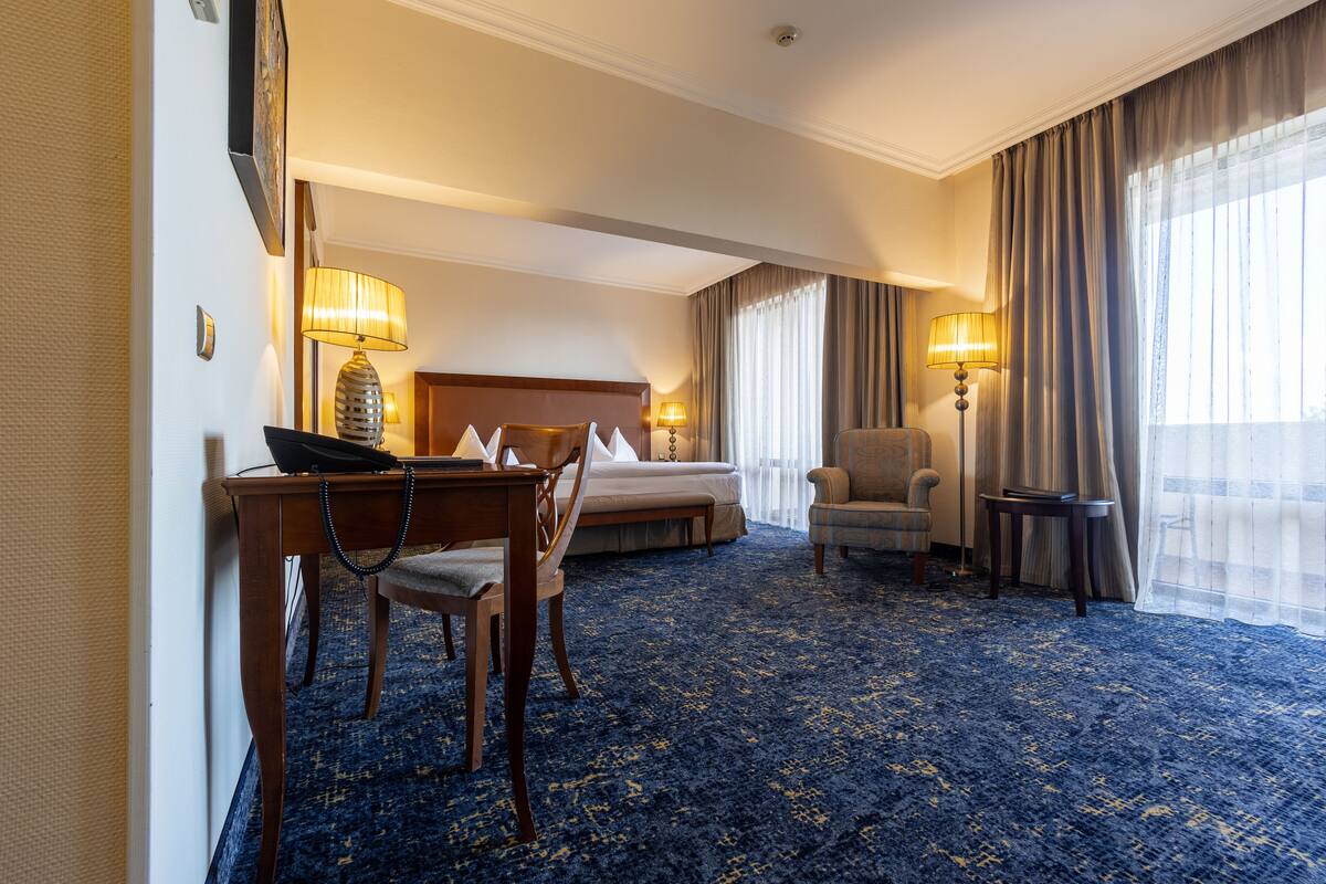 A hotel room with a bed, a desk with a telephone, two chairs, a small table, and a lamp.