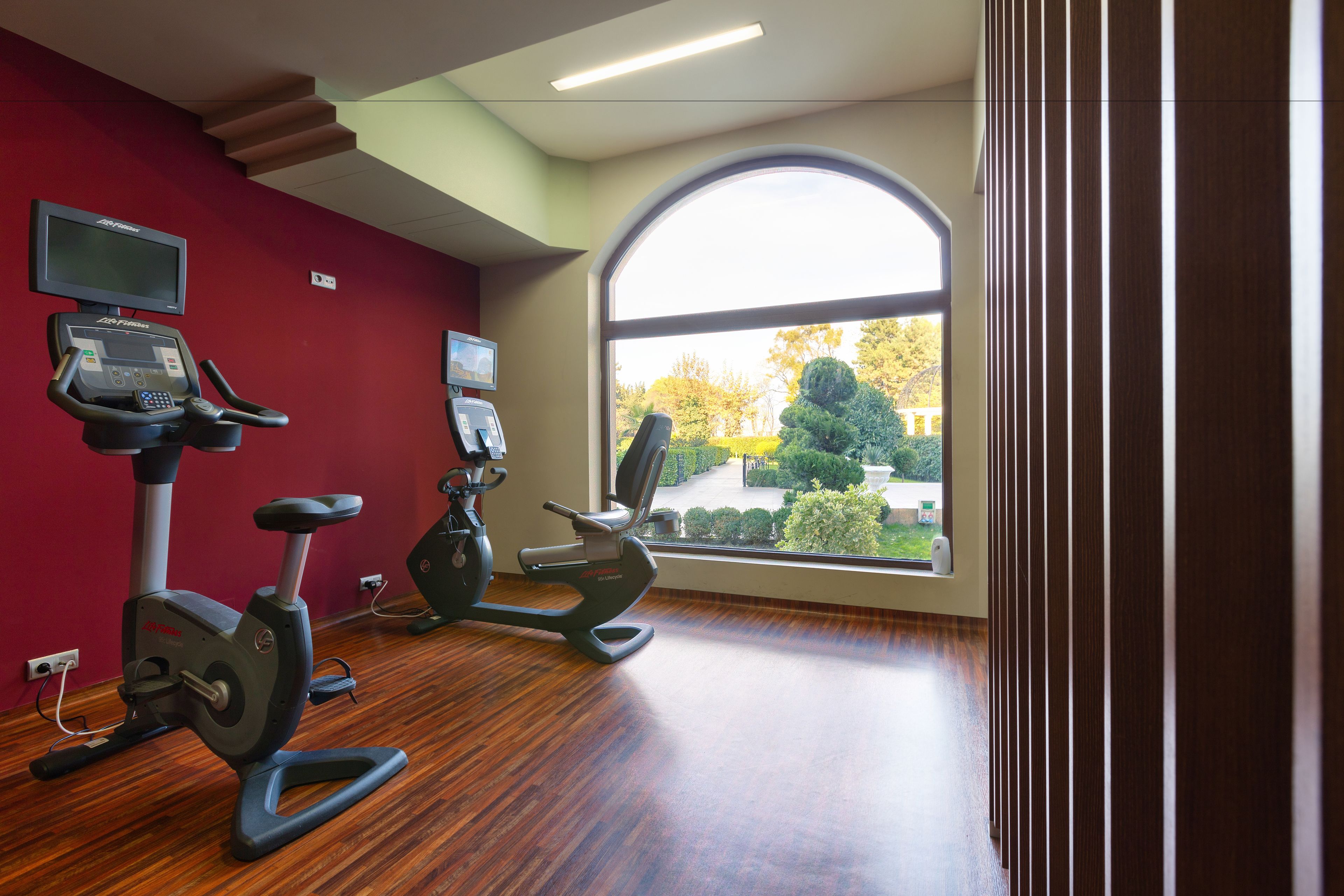 A gym with two stationary bikes, a TV on the wall, and a large window with a view of trees and a pathway.