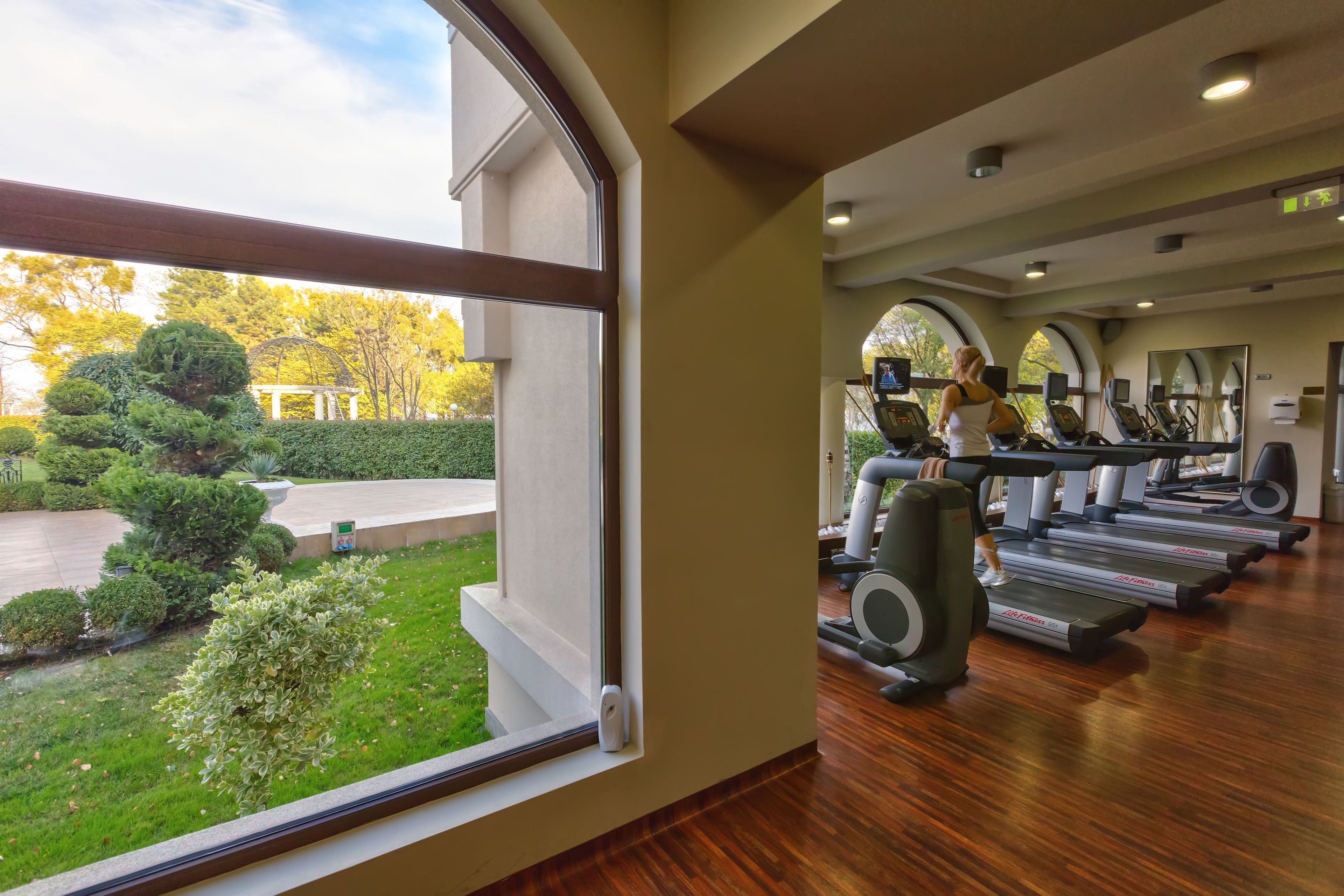 A gym with treadmills and exercise equipment, large windows with a view of a garden and trees.