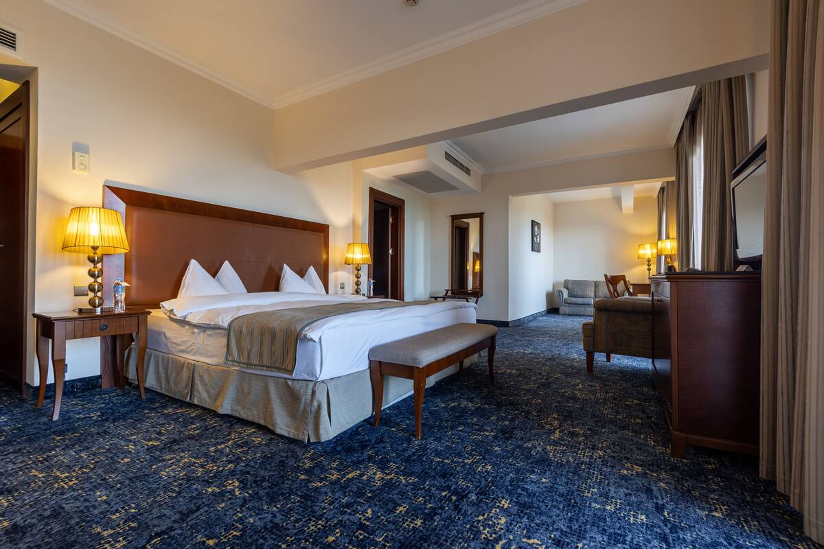 A hotel room with a large bed, bedside tables, a bench, a chair, and a desk.