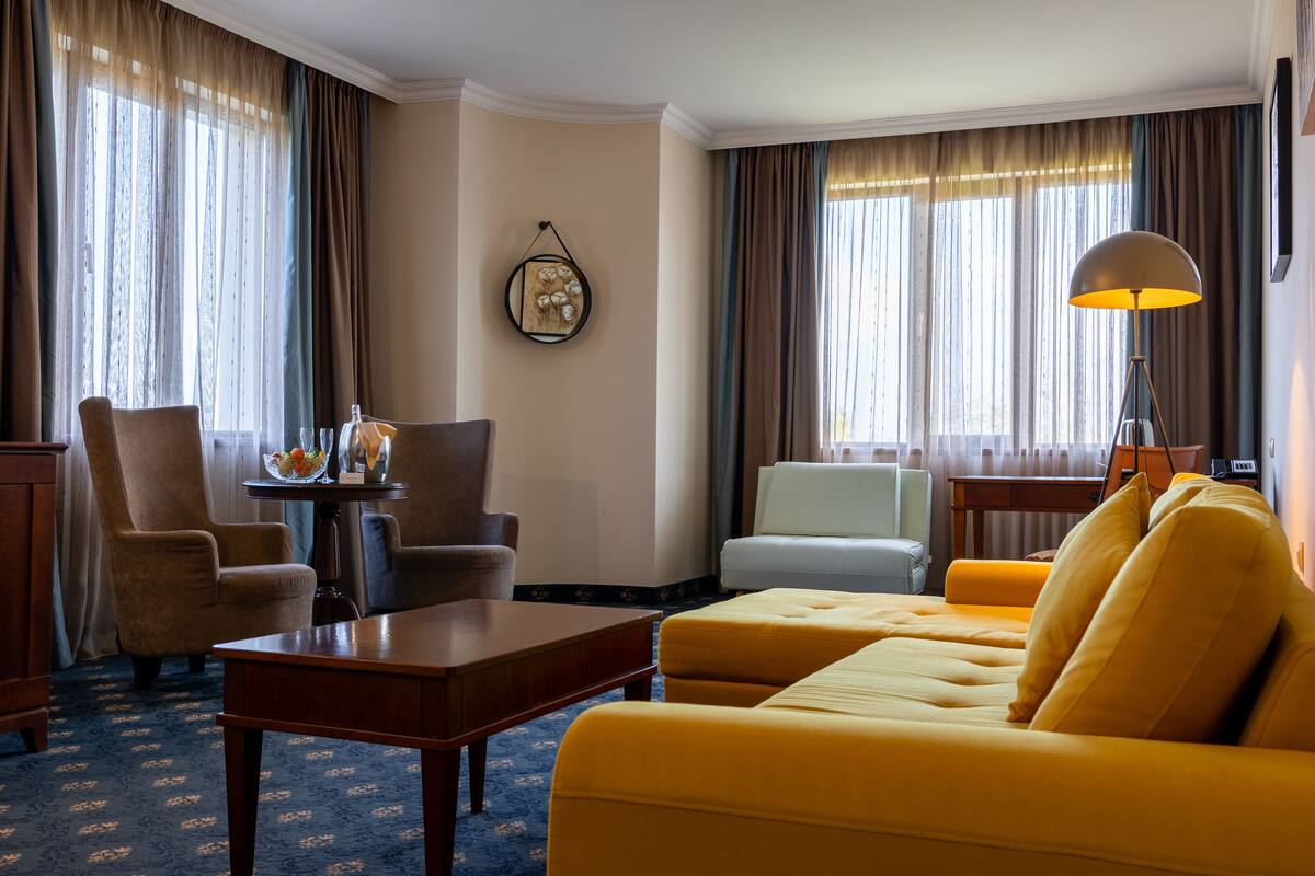 A hotel room with a yellow sofa, a wooden coffee table, two armchairs, a small table with a fruit bowl, and a lamp.