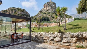 A patio with stone walls and a glass enclosure, overlooking a mountainous landscape with a white fence and palm trees. - Baglio dello Zingaro (Castellammare del Golfo)