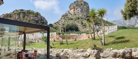A patio with stone walls and a glass enclosure, overlooking a mountainous landscape with a white fence and palm trees.