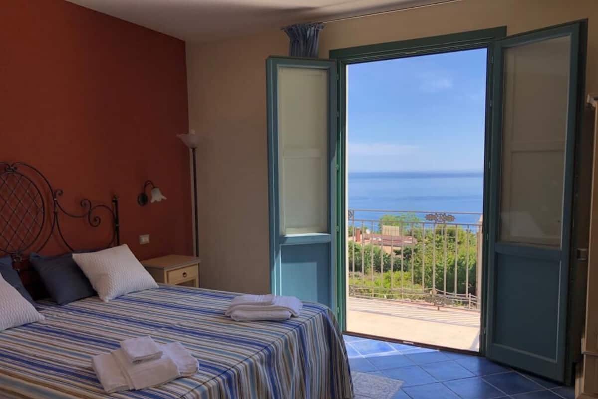 Un dormitorio con una cama, una mesita de noche y vista al mar a través de puertas abiertas.