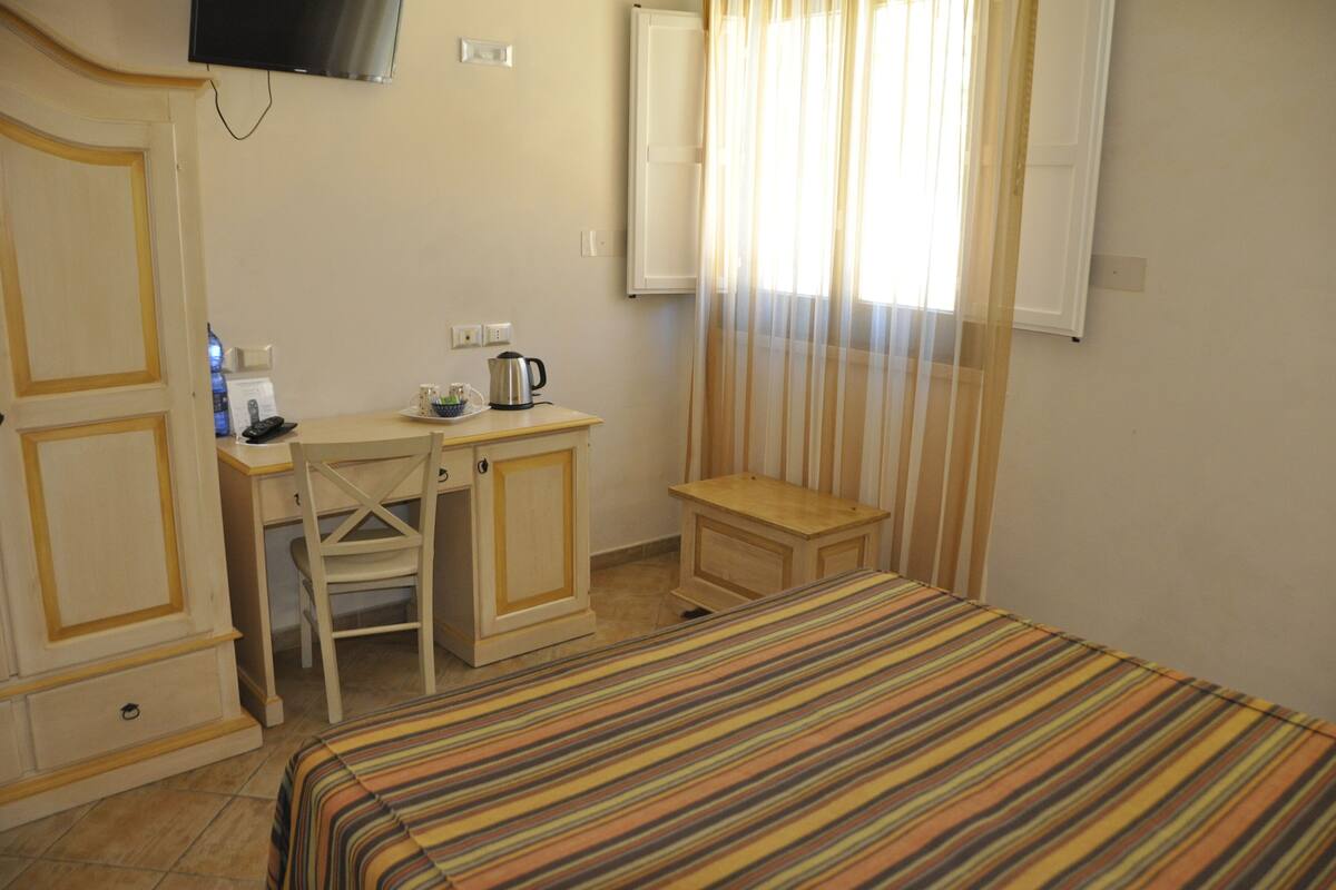 Habitación de hotel con cama, un escritorio con silla, una mesita, televisor montado en la pared y ventana con cortinas.
