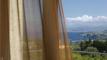 A view through a window with curtains, featuring a coastal landscape with mountains, a body of water, and a distant city.