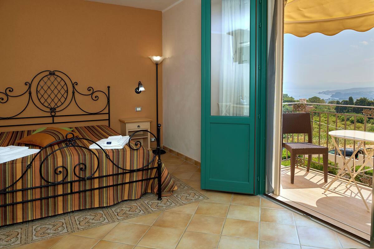 Habitación de hotel con cama, mesita de noche, lámpara y vistas al exterior.