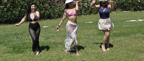 Three people dancing in a grassy outdoor area with a hedge and flowers in the background.