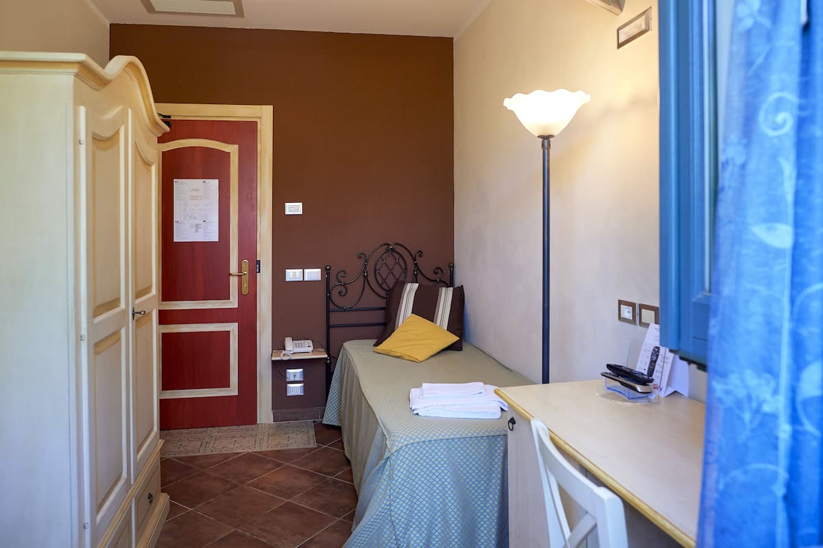 Una habitación pequeña y funcional con cama, escritorio y lámpara. Se observa una puerta con un aviso, y una mesita con teléfono y documentos.