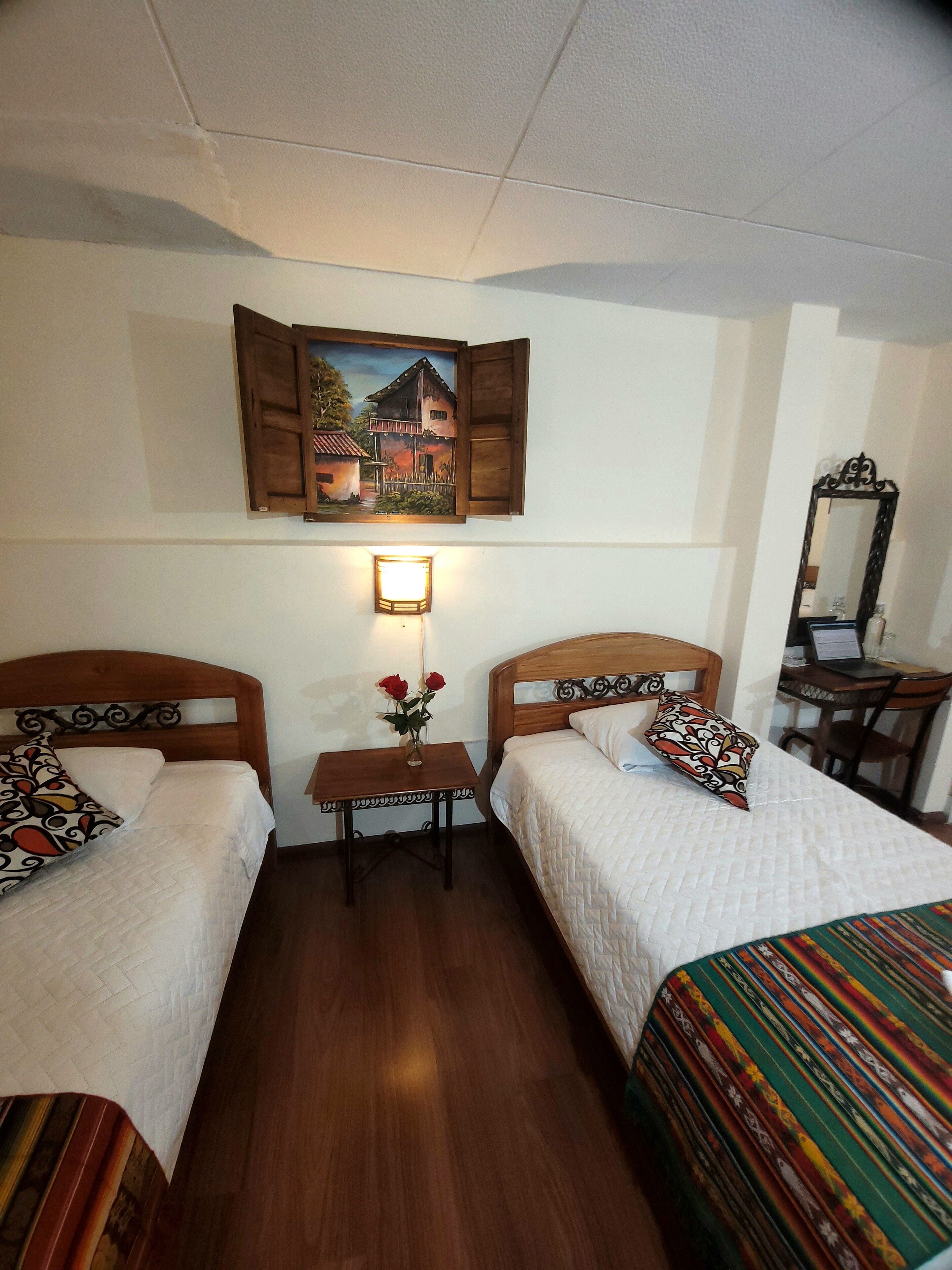 A room with two beds, a small table with a vase of flowers, a wall-mounted painting, and a mirror.
