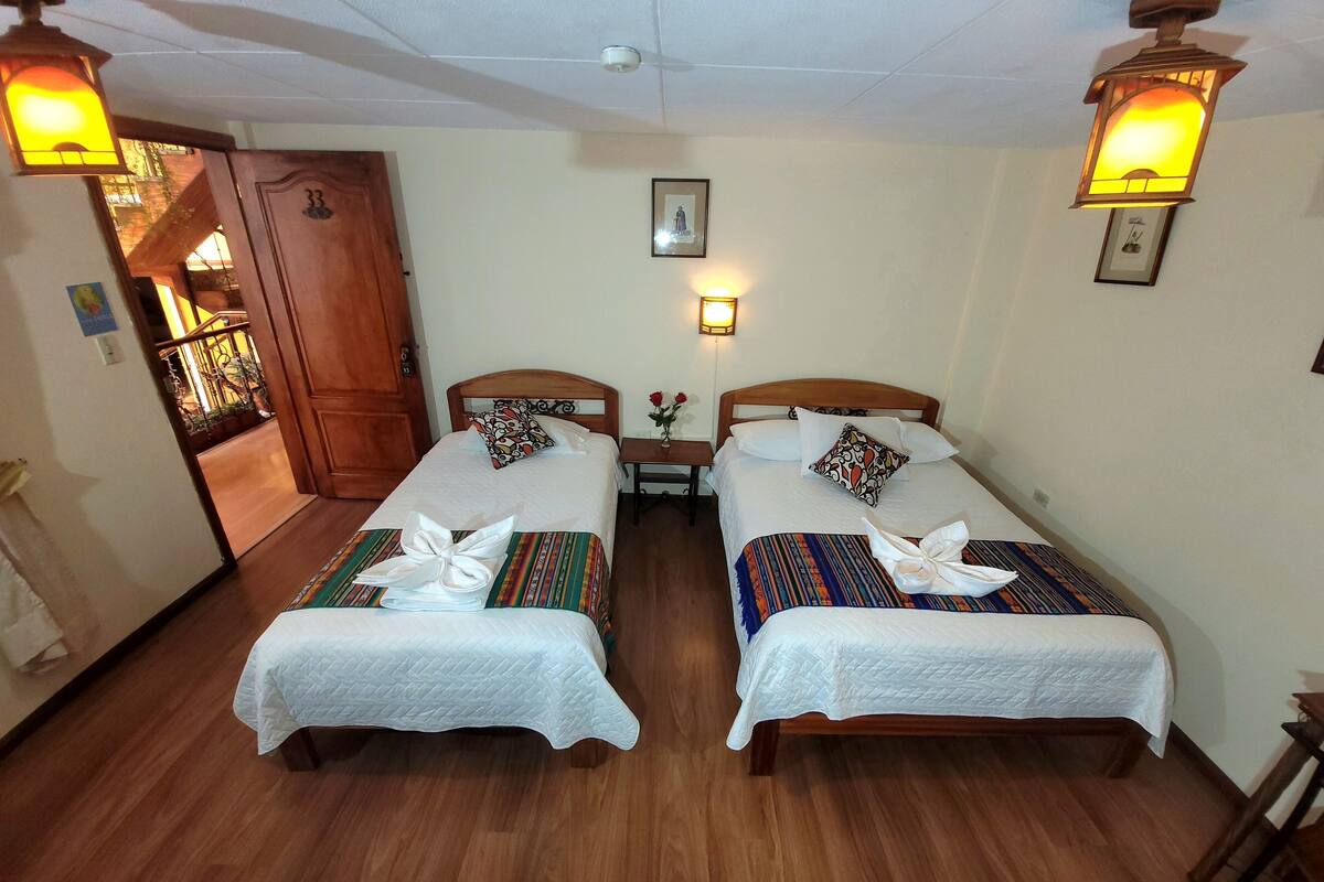 A hotel room with two single beds, wooden flooring, a wooden door, a small table with a vase of flowers, and wall-mounted lights.