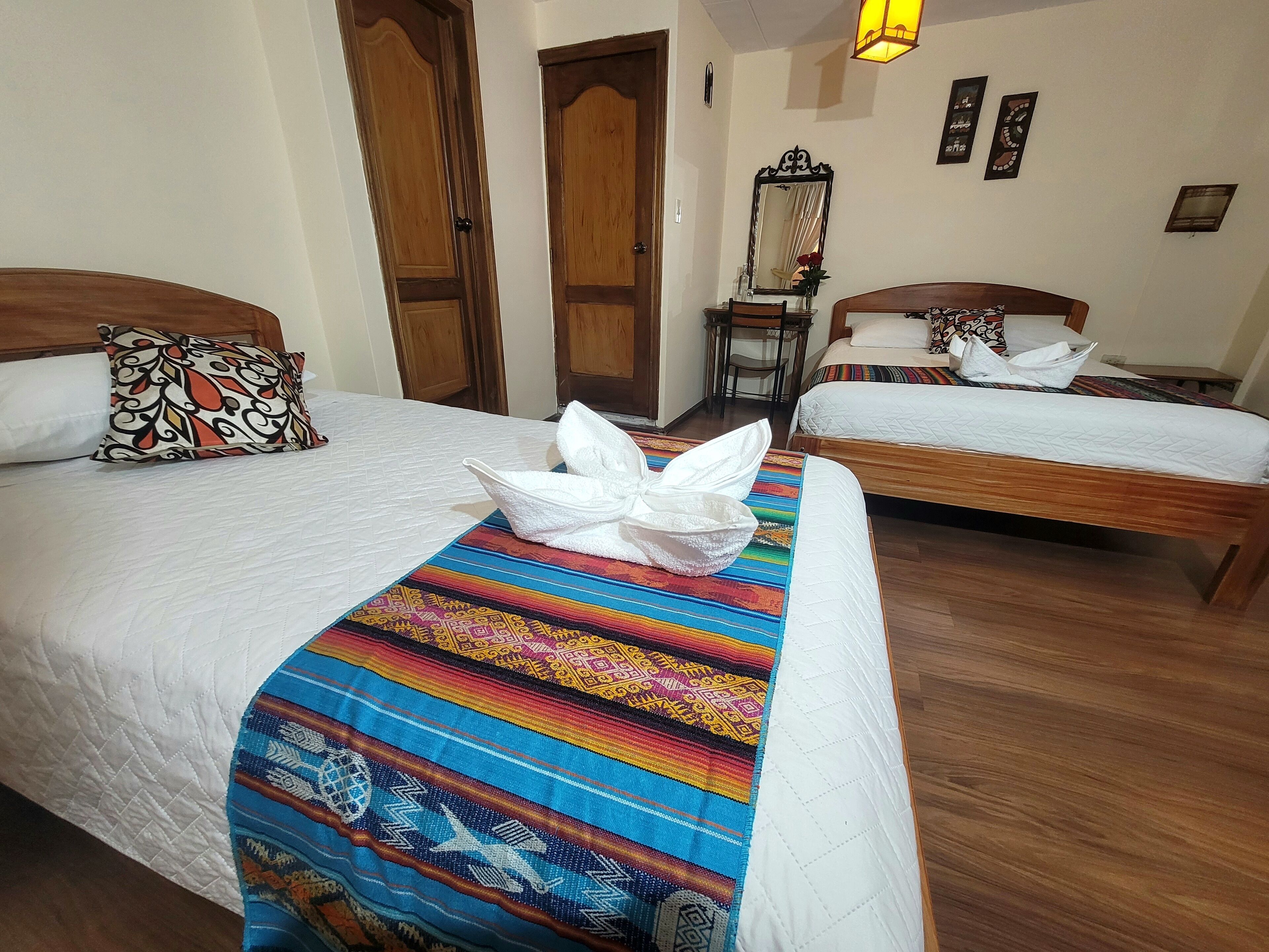 Une chambre d’hôtel avec deux lits, des têtes de lit en bois, du linge de lit blanc, une couverture colorée, un miroir et une petite table avec une chaise.