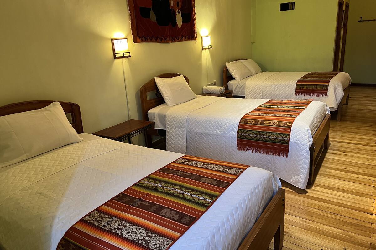 A hotel room with three beds, each with a white quilt and a patterned blanket. There are two wall-mounted lights and a tapestry on the wall.