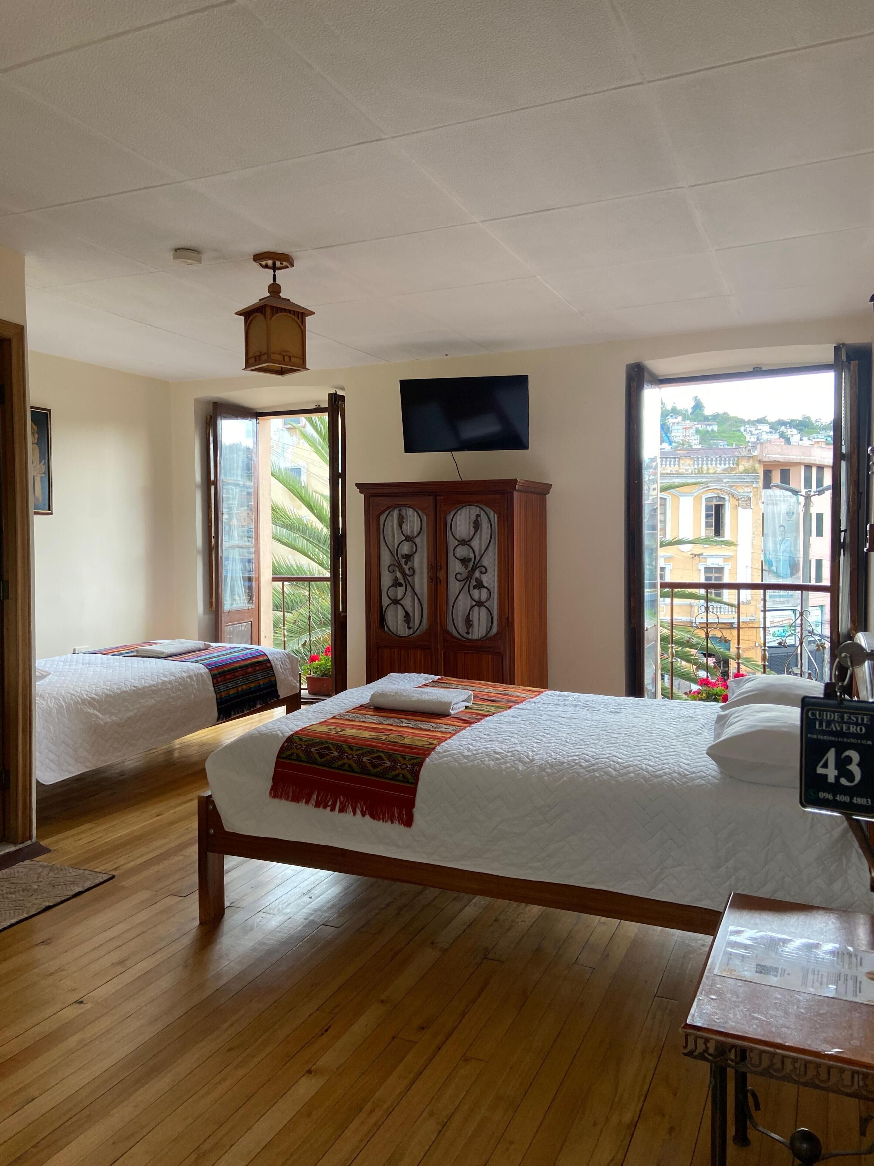 Een hotelkamer met twee bedden, een houten kast, een televisie en uitzicht op gebouwen door een groot raam.