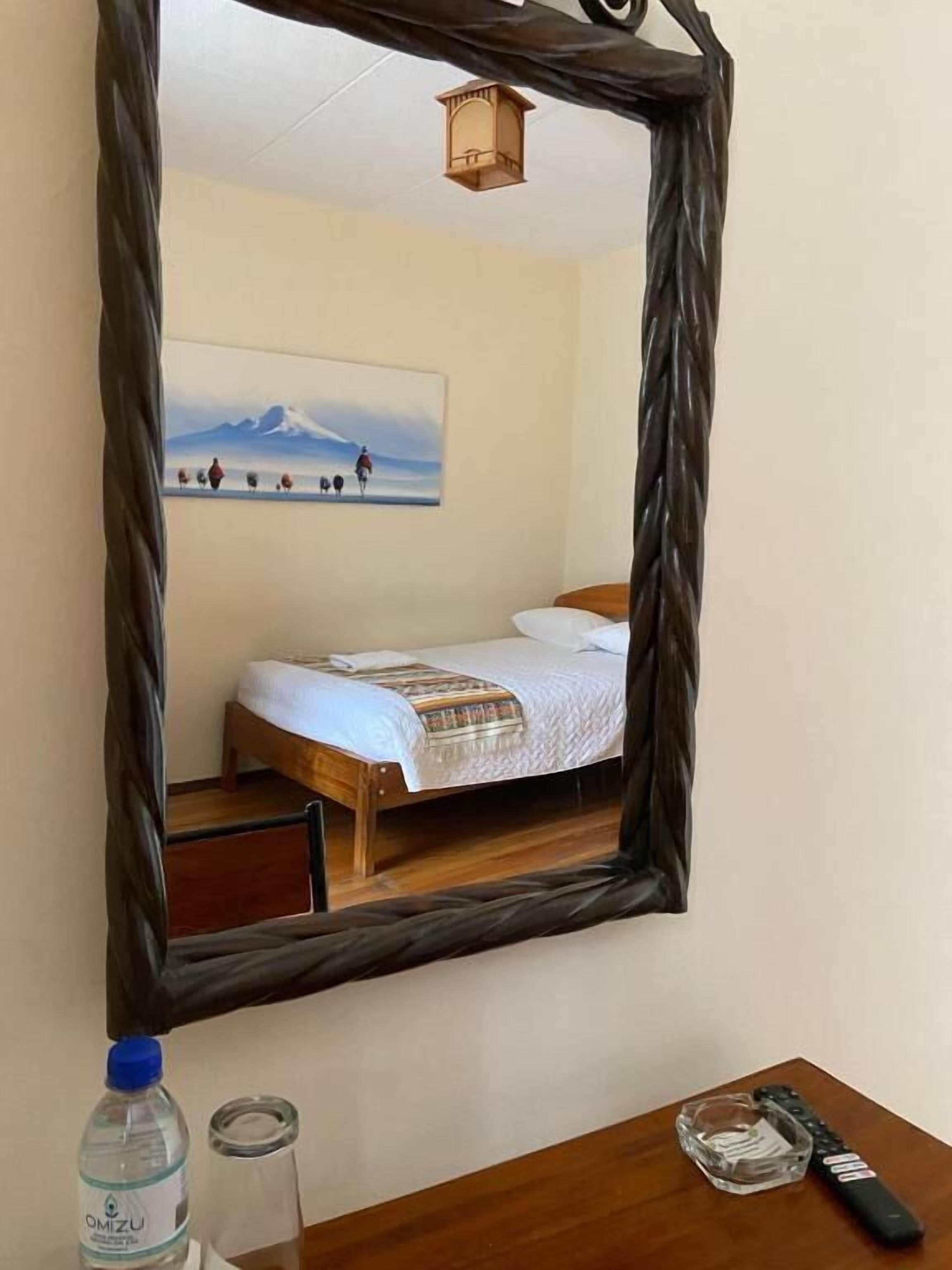 Una habitación con una cama, un espejo, una mesa de madera con un control remoto, una botella de agua y un vaso.
