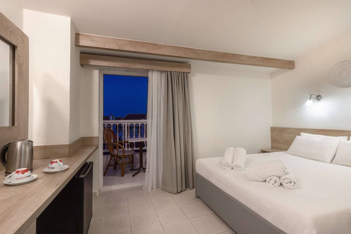 A hotel room with a bed, a desk with a kettle, a chair, and a balcony with a view of the evening sky.