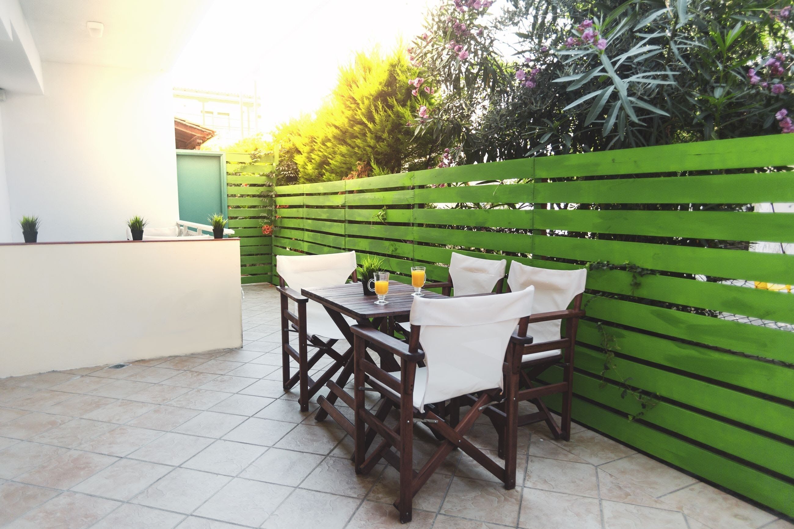 Eine Terrasse mit Holztisch und -stühlen, einem grünen Zaun und Kübelpflanzen.