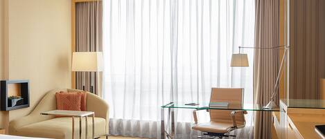 A hotel room with a bed, a desk, a chair, a lamp, and a large window with curtains.