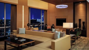 Habitación de hotel moderna con un sofá grande, una mesa de centro y una televisión. Amplias ventanas ofrecen vistas del horizonte de la ciudad.