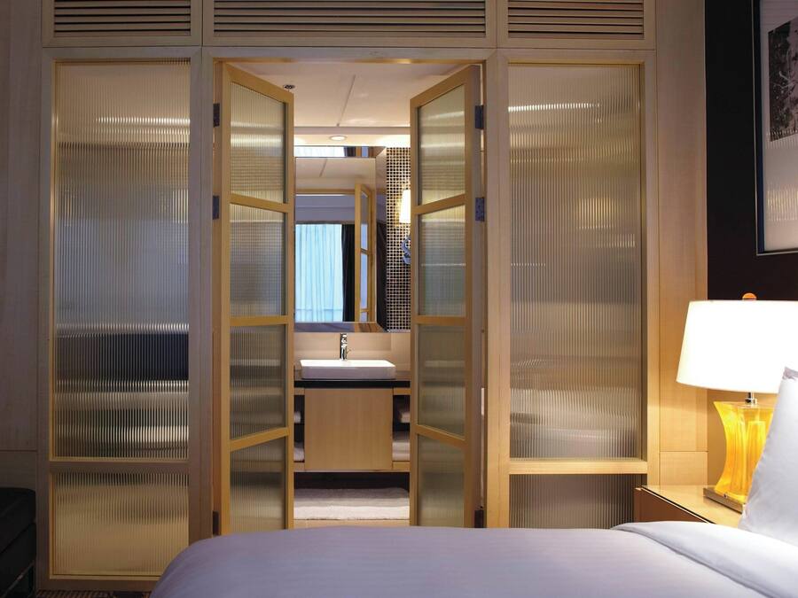 Habitación de hotel con cama, mesita de noche con lámpara, baño con lavamanos y espejo, y armario con puertas correderas.