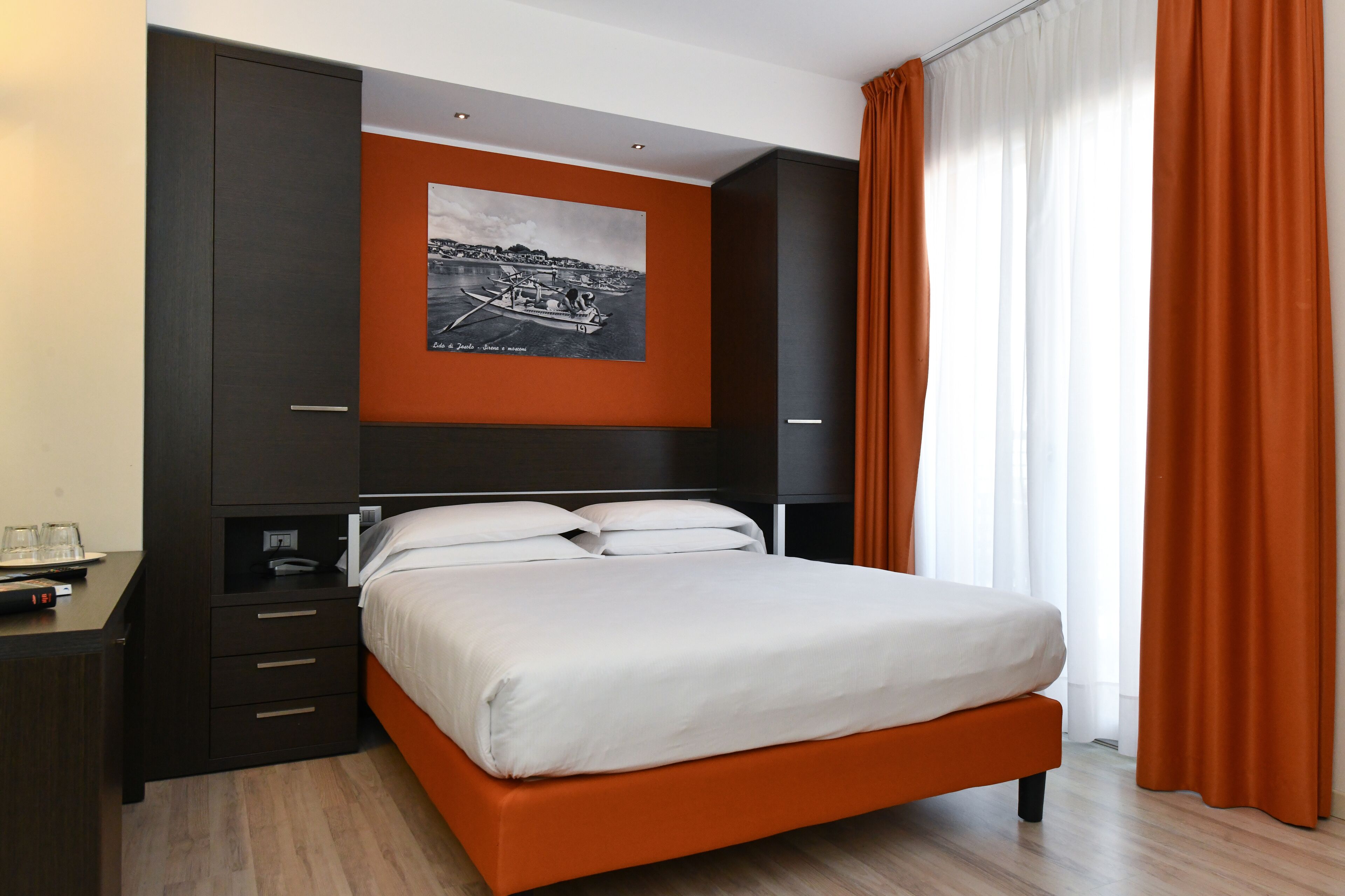 A modern hotel room with a large bed, a nightstand, a desk, and a wall-mounted picture.