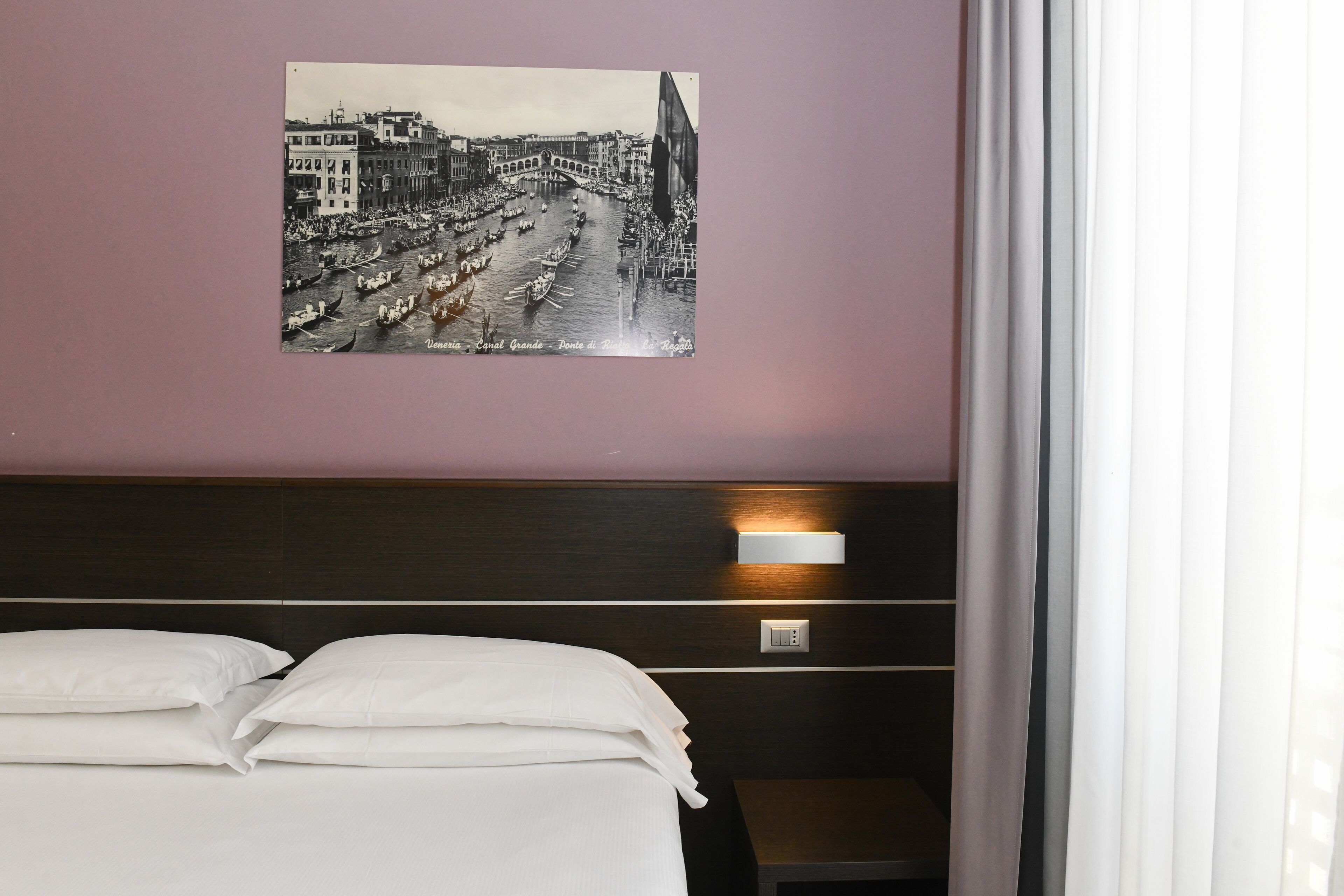 Habitació d'hotel amb un llit, una tauleta de nit, una làmpada penjada a la paret i un quadre amb una vista de canal.