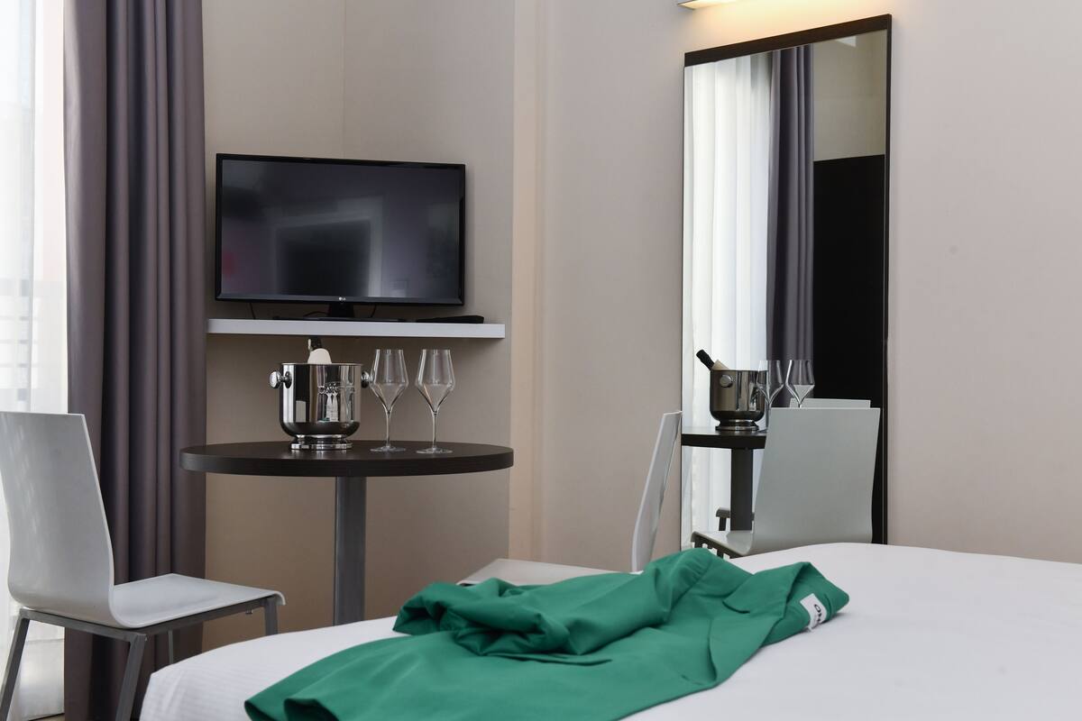 A hotel room with a bed, a TV on a stand, a table with a pitcher and glasses, and a mirror.