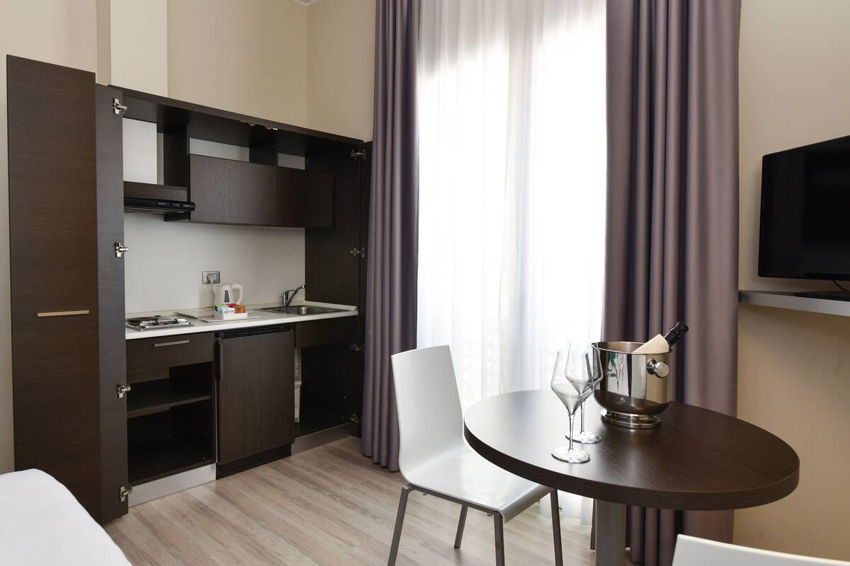 A compact hotel room with a kitchenette, a dining table with a chair, a TV mounted on the wall, and a window with curtains.