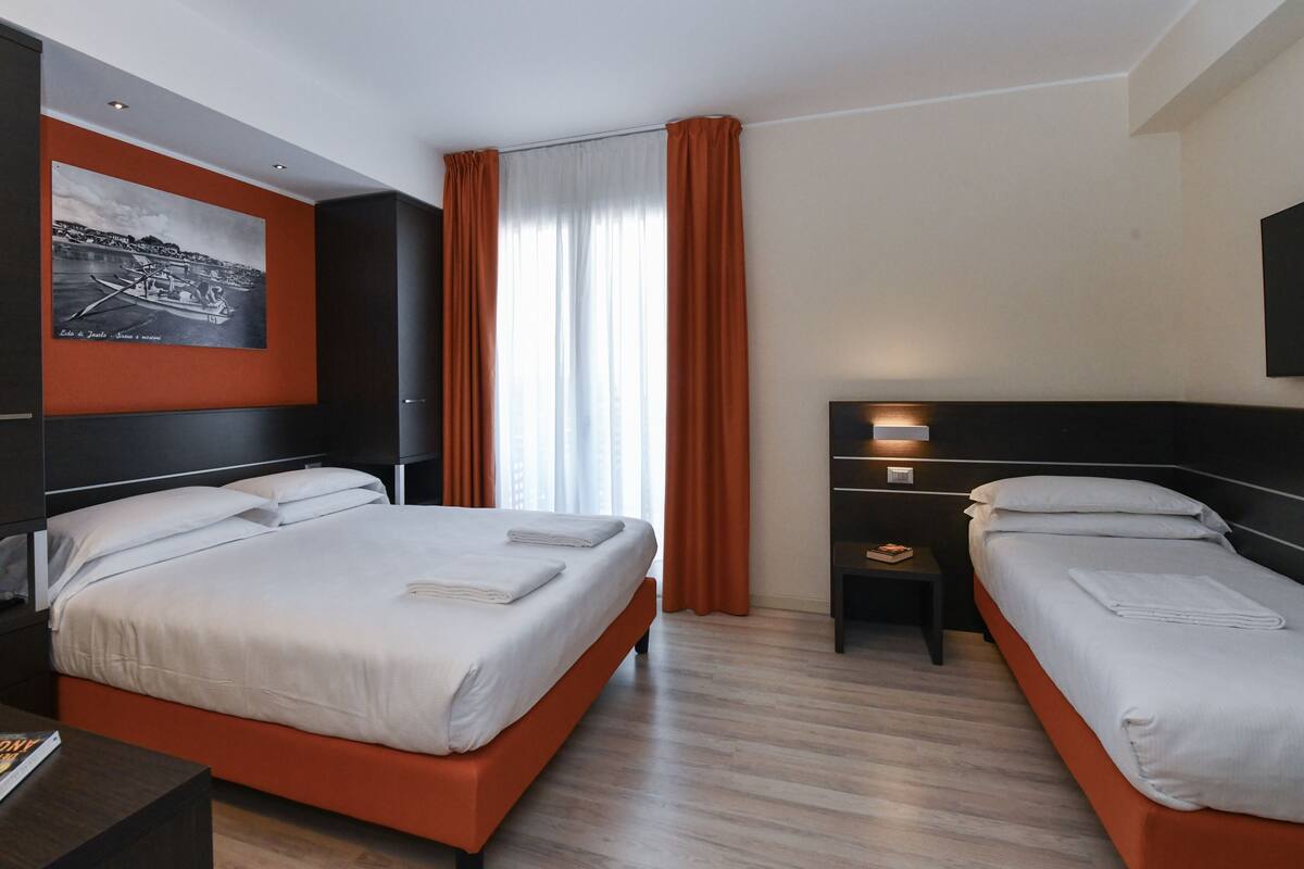 A hotel room with two beds, a large window with curtains, a painting on the wall, and a bedside table with a lamp.
