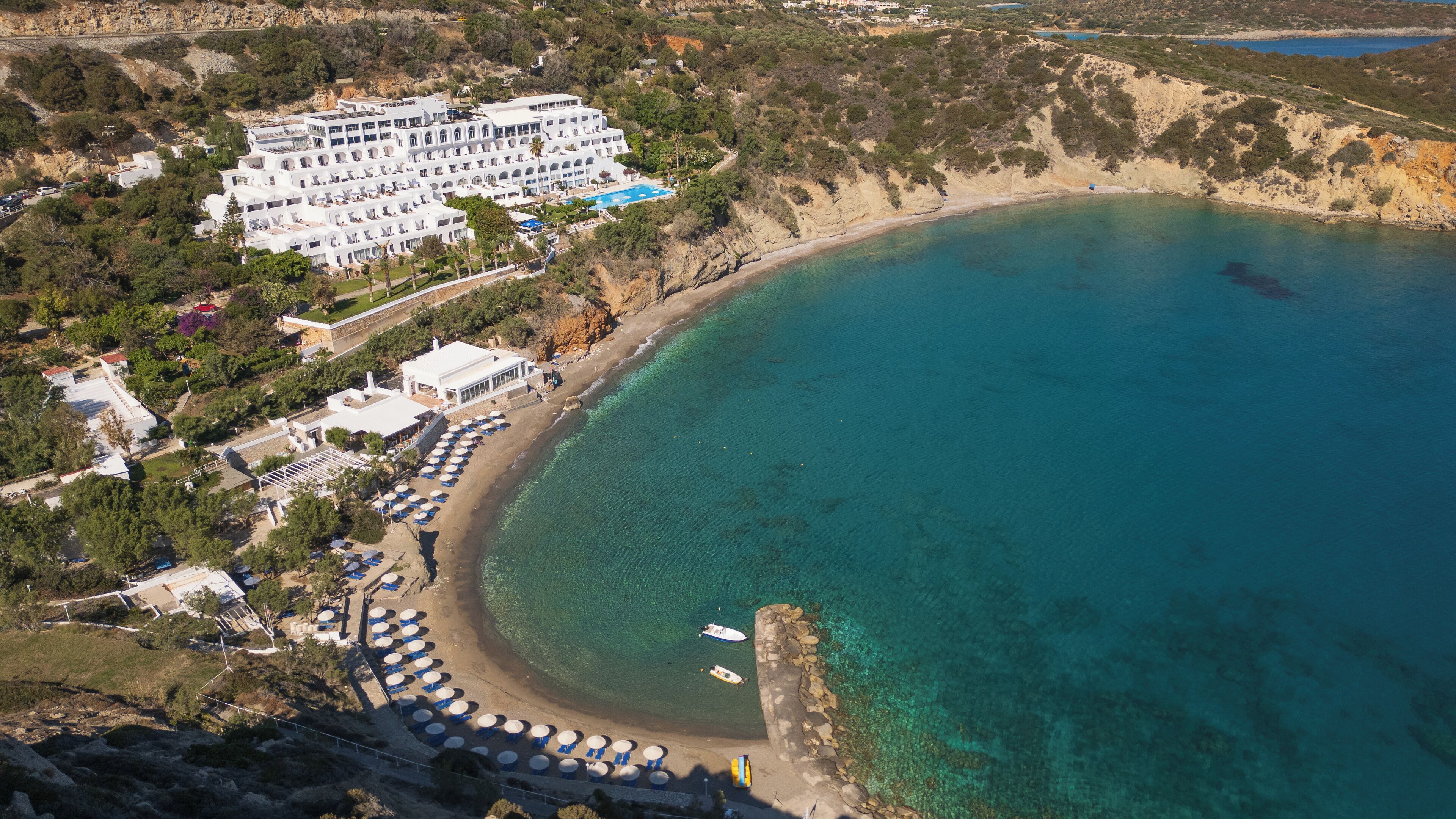 A coastal hotel with multiple white buildings, a beach with umbrellas, and clear blue waters.